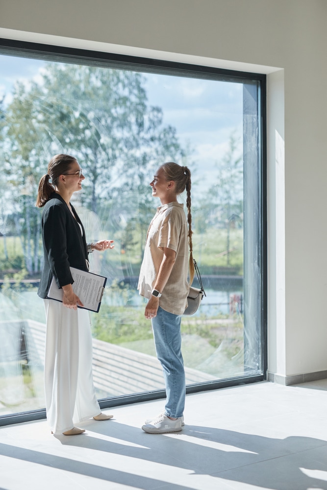 Agente immobilière guide une cliente lors d'une visite de maison. Agente immobilière et cliente souriantes discutent lors d'une visite. Grande fenêtre panoramique, intérieur moderne et vue nature.